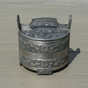 以下が含まれることがあります： 蓋と2つのハンドルが付いた、銀色の丸い容器。容器は花柄とスクロールワークパターンで装飾されています。蓋には装飾的な花の模様があります。容器は木製の表面に置かれています。