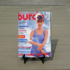 May include: A vintage fashion magazine titled "Burda" is displayed on a black stand. The cover features a woman in a blue patterned outfit. The magazine's title is in large red letters, with additional text in various colors. The magazine is dated 4/96.