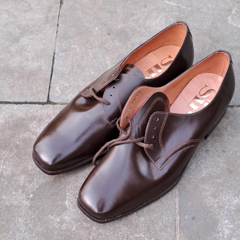 May include: A pair of brown leather dress shoes with a square toe and lace-up closure. The shoes have a glossy finish and are set on a gray stone surface. The interior of the shoe is a lighter brown color.