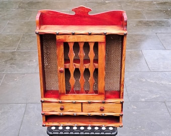 May include: A small, wooden cabinet with a red top and trim, featuring a central door with a decorative cutout design and two small drawers. The cabinet has a woven backing and is adorned with small, dark studs.