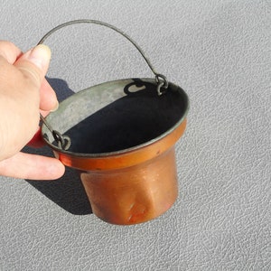 May include: A small, antique copper bucket with a metal handle. The bucket has a cylindrical body with a slightly flared rim and a tarnished, aged appearance. The handle is made of a thin, curved metal wire.