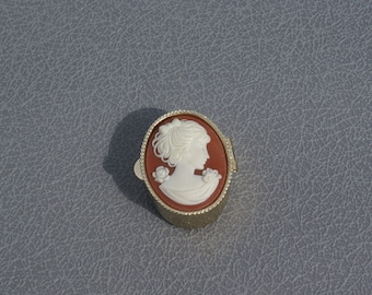 May include: Oval cameo brooch with a white profile on a reddish-brown background, in a gold-toned frame. The cameo shows a woman with flowers. The brooch is on a grey surface.