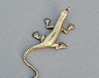 May include: A gold-colored lizard brooch with detailed scales and a curved tail. The reptile-themed jewelry piece is shown on a gray surface. The lizard's legs are outstretched, and its head is slightly raised.