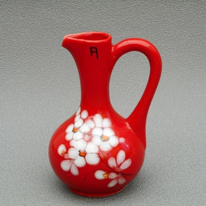 May include: A vibrant red ceramic pitcher with a curved handle and a spout. The pitcher is adorned with white floral designs with orange centers. The letter "A" is visible on the pitcher. Decorative home decor.