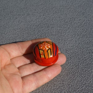 Op de afbeelding: Een kleine, ronde, rode houten doos met een deksel. Het deksel is versierd met een geometrisch ontwerp in oranje, geel en wit. De doos wordt in een hand gehouden tegen een grijze achtergrond.
