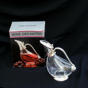 Könnte beinhalten: Eine klare Weindekanter mit silberfarbenem Griff und Boden. Die Dekanter steht neben der Originalverpackung mit der Aufschrift "WINE DECANTER". Die Dekanter hat eine einzigartige Form und ein einzigartiges Design.