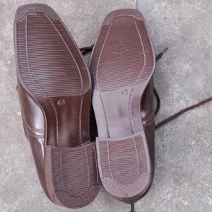 May include: A pair of brown leather dress shoes, soles facing up. The shoes have a square toe design and are size 43. The soles have a textured pattern for grip. Brown laces are visible.