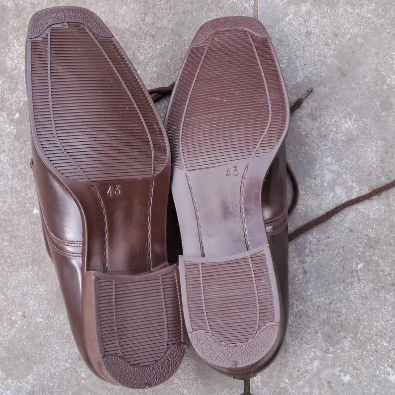 May include: A pair of brown leather dress shoes, soles facing up. The shoes have a square toe design and are size 43. The soles have a textured pattern for grip. Brown laces are visible.