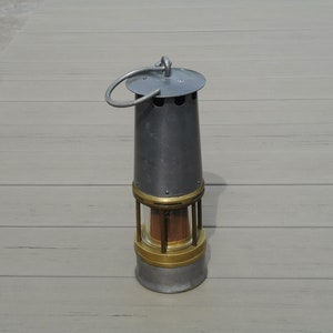 May include: A vintage miner's lamp with a cylindrical metal body, brass accents, and a glass enclosure. The lamp has a handle and a circular top. The lamp is gray and gold in color.