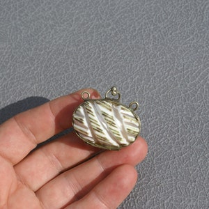 May include: An oval pendant with a silver-colored frame and a white and gold striped design. The pendant has two small loops on the sides and one on top for attaching to a chain. The pendant is held in a hand.