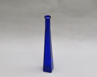 May include: A tall, cobalt blue glass bottle with a narrow neck and a square base. The bottle has a geometric, tapered design, and stands upright against a neutral background. The bottle's height is approximately 10 inches.