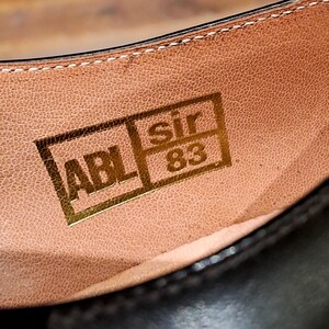 May include: Close-up of the inside of a brown leather shoe. The shoe's insole features a gold rectangular stamp with the letters "ABL", the word "sir", and the number "83". The stitching is visible along the top edge.