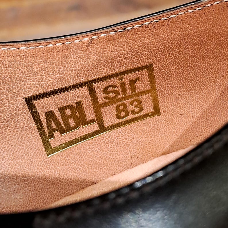 May include: Close-up of the inside of a brown leather shoe. The shoe's insole features a gold rectangular stamp with the letters "ABL", the word "sir", and the number "83". The stitching is visible along the top edge.