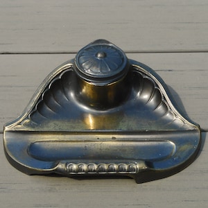 Peut inclure: Encrier ancien avec un design décoratif à festons. L'encrier est en métal avec un aspect vieilli et terni. Il comporte un petit couvercle rond avec un motif floral et un porte-plume intégré.