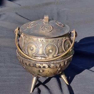 May include: A vintage metal pot with a lid, handle, and three legs. The pot is adorned with circular and crescent-shaped patterns. The lid has a small handle. The pot has a tarnished, antique appearance.