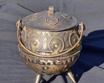 May include: A vintage metal pot with a lid, handle, and three legs. The pot is adorned with circular and crescent-shaped patterns. The lid has a small handle. The pot has a tarnished, antique appearance.
