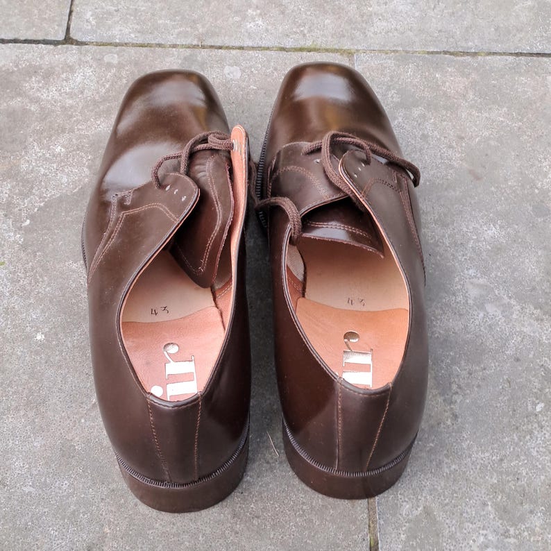 May include: A pair of brown leather dress shoes with laces. The shoes have a classic design with a rounded toe and a low heel. The interior of the shoe is visible, showing a light brown insole with the brand logo.