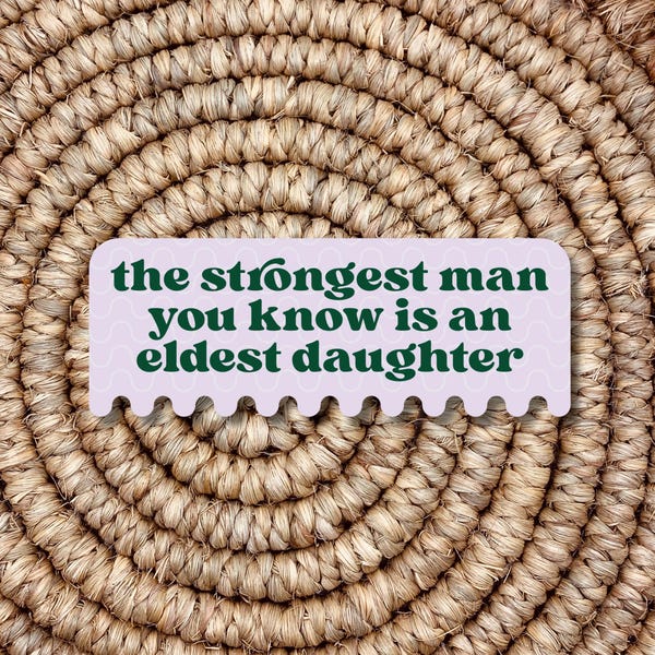 May include: A light purple sticker with a wavy border and dark green text that reads "the strongest man you know is an eldest daughter". The sticker is placed on a woven, circular, brown background.