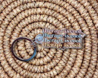 May include: A clear keychain with the text "therapy is cool" repeated four times in pink and blue lettering. The keychain is on a woven straw background.
