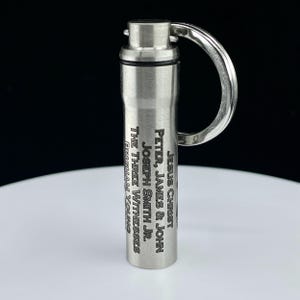 May include: A cylindrical silver-toned metal keychain pill container with a screw-top lid and a split ring. The side is engraved with the text: JESUS CHRIST, PETER, JAMES & JOHN, JOSEPH SMITH JR., THE THREE WITNESSES, BRIGHAM YOUNG.