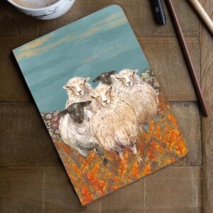 May include: A notebook featuring a sheep illustration, alongside a cup of coffee, ink, and pens, arranged on a wooden surface. The notebook depicts sheep in a field with a blue sky. The coffee cup is white with a dark rim.