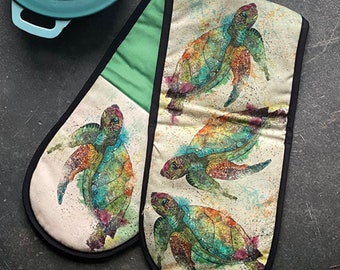 May include: A pair of green and cream oven mitts with a watercolor design of four colorful sea turtles. The mitts have black trim.
