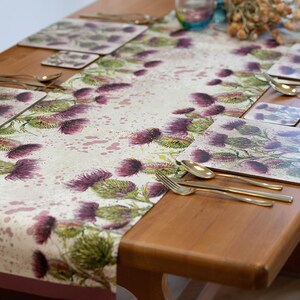 Può includere: Una mise en place con un runner da tavolo floreale rosa e verde con un motivo a cardo. Il runner è su un tavolo di legno con posate dorate e tovagliette abbinate.