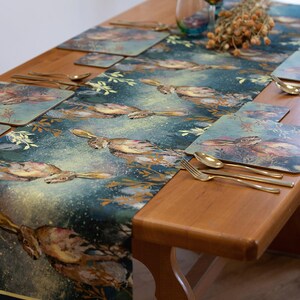 Può includere: Un runner da tavolo blu e oro con un motivo di lepri in un ambiente boschivo. Il runner è su un tavolo di legno con posate dorate e sottopiatti.