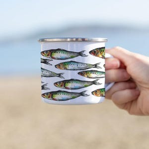 May include: White enamel mug with a pattern of colorful sardines. The mug is being held by a hand against a blurred background of a beach and ocean.