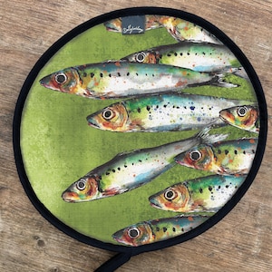 Könnte beinhalten: Runder Topflappen mit schwarzem Rand und Schlaufe. Er zeigt eine Aquarellillustration von Sardinen in Grün-, Blau-, Gelb- und Orangetönen. Der Markenname "Dollyhotdog" ist oben aufgedruckt.