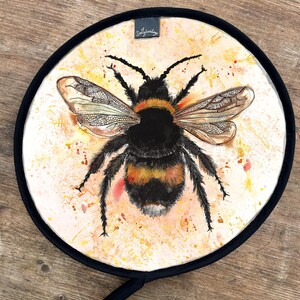 Peut inclure: Un dessous de plat rond avec une illustration de bourdon à l'aquarelle. L'abeille est noire et jaune avec des ailes détaillées, sur un fond clair et moucheté. Une bordure noire et une petite étiquette avec du texte complètent le design.
