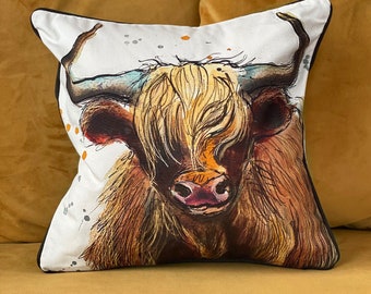 May include: A white throw pillow with a watercolor illustration of a brown and white Highland cow with long hair. The cow's face is the focus of the image, and its eyes are closed. The background is white with small orange and brown dots.