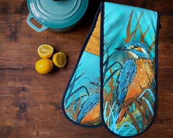 May include: A pair of blue and yellow oven mitts with a bird design. The mitts are made of fabric and have a dark blue trim. The bird is a kingfisher, and it is perched on a branch with green leaves. The background is a light blue color.