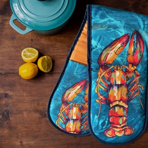 May include: A pair of blue oven mitts with a lobster design. The lobsters are painted in bright orange, red, and yellow colours. The mitts are on a wooden surface.