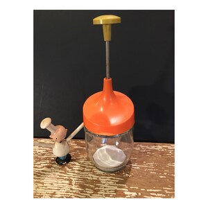 May include: Orange and yellow vintage hand-crank food chopper with a glass jar and a clear plastic lid. The chopper is on a wooden surface.