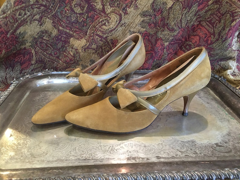 May include: A pair of tan suede high heels with a silver trim and a bow detail. The shoes have a pointed toe and a stiletto heel.