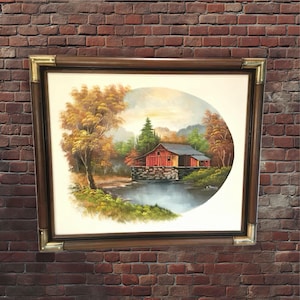 Puede incluir: Una pintura enmarcada de un granero rojo en un paisaje rural. El granero está rodeado de árboles y un río, y el cielo es de un azul suave con nubes. La pintura está en un marco marrón con detalles dorados.
