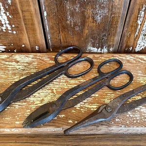 May include: Three antique metal shears with large finger loops rest on a weathered wooden surface. The shears have a dark, aged appearance, suggesting they are vintage tools. The wooden surface has a distressed look, adding to the rustic aesthetic.