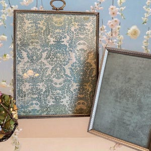 Può includere: Due cornici rettangolari vintage. Una cornice presenta un motivo damascato verde acqua e crema, l'altra ha uno sfondo verde acqua pieno. Entrambe le cornici hanno bordi metallici ornati. Una composizione floreale decorativa è in primo piano.