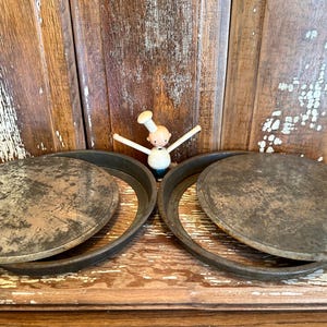 Puede incluir: Dos moldes redondos de metal vintage para pasteles con tapas, que muestran signos de desgaste. Una figura de chef de madera se encuentra entre los moldes. Los moldes están sobre una superficie de madera, con un fondo de madera rústica.