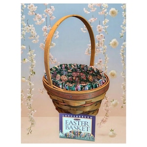 May include: A round woven wooden basket with a tall arched handle and a ruffled fabric liner featuring a colorful egg pattern. A rectangular card in front displays the text Longaberger Easter Basket with an illustration of decorated eggs.