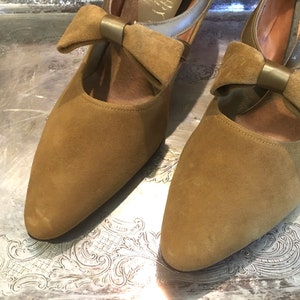 May include: A pair of brown suede heeled shoes with a bow detail on the front. The bow has a gold metal accent.