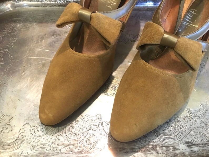 May include: A pair of brown suede heeled shoes with a bow detail on the front. The bow has a gold metal accent.