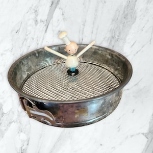 May include: A vintage metal cake pan with a wire mesh bottom and a hinged side. A small, wooden chef figurine with outstretched arms stands in the center. The chef wears a white uniform and hat, with a black bow tie.
