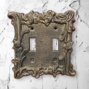 May include: A double light switch wall plate featuring a metallic bronze finish with a textured, hammered center and a raised, ornate border decorated with sculpted floral and leaf motifs, including four screw holes for installation.