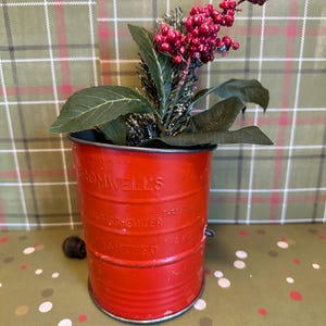 Op de afbeelding: Een rode metalen emmer met de tekst "Cromwell's" en "Guaranteed" in reliëf. De emmer bevat een decoratief arrangement van groene bladeren, kunstmatige dennentakken en rode bessen. De emmer staat op een groen-wit gestippeld oppervlak.