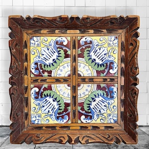 May include: Square wooden frame with ornate carvings, holding four decorative ceramic tiles. The tiles feature a floral design with blue, yellow, green, and red colors. The frame is a warm brown color.