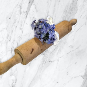 May include: A wooden rolling pin with a light brown finish and a natural wood handle. The rolling pin is decorated with a bouquet of purple flowers.