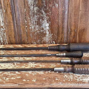 May include: Four vintage screwdrivers with wooden handles and metal shafts are arranged on a weathered wooden surface. The tools vary in size and handle design, showcasing a rustic aesthetic.