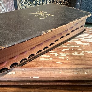 Puede incluir: Un libro vintage encuadernado en cuero negro con detalles dorados y un dise&ntilde;o decorativo en la portada. El libro est&aacute; cerrado y la encuadernaci&oacute;n es visible, mostrando que las p&aacute;ginas est&aacute;n cosidas juntas. El libro descansa sobre una superficie de madera.